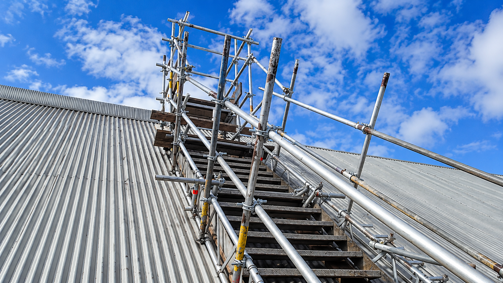 Andaime tubo roll montado em telhado de telha metalica de galpao industrial, sob ceu azul, para servico em cobertura industrial